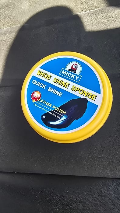 The image displays a circular yellow container of shoe polish, featuring a blue and white label. The brand name "MICKY" is prominently visible on the label, accompanied by a cartoon character. Further text on the label identifies the product as a "SHOE SHINE SPONGE" for "QUICK SHINE" and "LEATHER POLISH," with the tagline "Cleans and Shines in One Easy Step." An illustration of a shiny black shoe is also depicted on the label, indicating the product's function. The container rests on a dark, textured surface, which shows lighter, granular areas in the background, suggesting a practical setting. No specific activity is occurring. This scene is situated in Shimbi Mashariki, Tanzania.