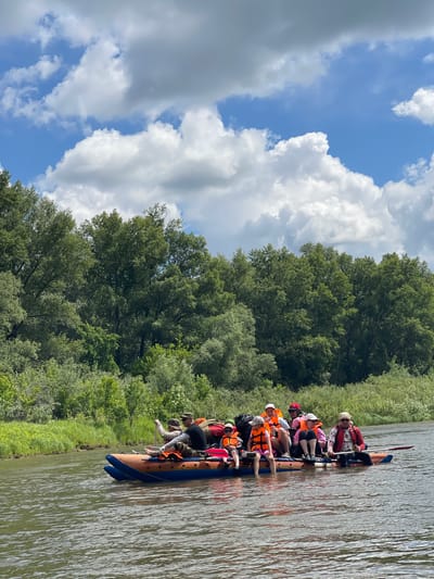 A group of ten people, including adults and children, are seated on an inflatable raft on a wide, calm river. The raft is orange and blue, with several bags strapped to its surface. The people are wearing casual clothing and life jackets in various colors, including orange, pink, and red. Some are holding paddles, and one person appears to be steering. The raft is positioned in the foreground, with the people looking in different directions. Behind the raft, dense green trees line the riverbank, forming a thick forest that extends to the horizon. The sky above is blue with scattered white, fluffy clouds. The overall scene depicts a leisurely boat trip on a sunny day in a natural environment, likely the area of Samorodovo, Russia, given the lush forest and river setting.