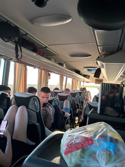 The image shows the interior of a passenger bus. People are seated in rows, looking at phones, using cameras, or resting. Overhead compartments contain bags and personal items. Windows are covered by curtains. The view outside shows a landscape suggesting a rural area, possibly near Bošnjaci, Croatia, given the query context.