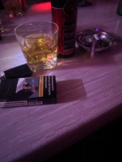 A close-up, high-angle image captures a collection of items on a light-colored wooden table within a dimly lit indoor establishment in Dolni Tsibar, Bulgaria. In the center-left, a clear glass tumbler holds an amber liquid, likely an alcoholic beverage, with ice cubes visible. Immediately behind the glass, a red "HELL ENERGY CLASSIC" beverage can stands upright. To the right of the energy drink can, a metallic ashtray contains several extinguished cigarette butts and ash, with one unlit cigarette horizontally resting across its rim. In the foreground, a black "DUNHILL" cigarette pack, bearing Bulgarian health warnings, is open on the table, partially covering a black cigarette lighter. The environment is characterized by low, artificial illumination, with prominent purple and red ambient lighting reflecting off the surfaces and casting shadows from the objects. No individuals are visible.