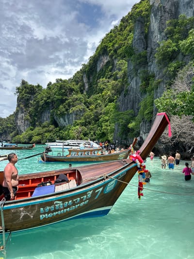 In Haad Hin Khom, Thailand, a body of clear, turquoise water is dotted with several wooden longtail boats. In the foreground, the bow of one such boat, adorned with a colorful garland of flowers and a pink ribbon, is prominently visible. A man in black shorts stands on the deck of this boat, looking to his left. Behind him, the interior of the boat reveals orange seats and a blue mat.
Further into the water, other longtail boats are anchored. One boat displays Thai script and the name "Piernnaree" in blue lettering. Several other boats, including a white motor yacht, are further out. A group of people, some wading in the water and others on boats, are visible in the midground. The background is dominated by a steep, lush green cliff face, characteristic of the region. The sky above is partly cloudy with a mix of grey and white clouds.