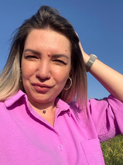 A woman is taking a selfie outdoors against a bright blue sky. She is wearing a pink button-up shirt and a necklace with a heart-shaped pendant. Her left hand is in her hair, and her right arm is raised behind her head, revealing a silver watch on her wrist. She has blonde and dark brown hair and is wearing hoop earrings. The background shows some green grass and foliage, suggesting a natural or park-like setting. There are no identifiable landmarks in the image that would indicate the specific location within Яново-Грушевский, Russia.