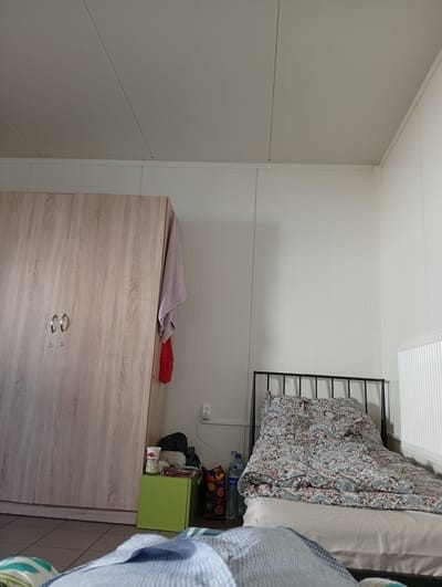 A bedroom scene is depicted with a wardrobe, a bed, and various objects. The room has white, paneled walls and a white, paneled ceiling. A large, light wood-grain wardrobe stands on the left side of the image. Two oval-shaped, silver handles and two smaller circular ones are visible on its doors. A light pink towel hangs from the top right corner of the wardrobe, with a red piece of fabric below it.

To the right of the wardrobe, near the corner, is a small, bright green cube-shaped piece of furniture. On top of it sits a white cup with red text and a picture of fruit, possibly yogurt or a similar product. Behind this cube, a black backpack is partially visible, along with a round, cream-colored object and a few books or binders. Further to the right, a patterned bag with fruit designs is placed on the floor. A bottle of water with a blue label is situated next to it.

The right side of the image features a black metal bed frame. The bed is covered with a comforter that has a complex floral pattern in shades of blue, green, and maroon on a light background. A radiator with vertical white fins is mounted on the wall to the far right. An electrical outlet with a single socket is visible on the wall above the radiator.

In the foreground, a portion of a bed with blue and white checkered bedding is visible, along with other bedding elements showing blue and white stripes, and green and white stripes.

The lighting suggests it is daytime, with soft, even illumination throughout the room. The overall impression is of a functional, perhaps somewhat cluttered, living space. No specific activity is clearly taking place. The location is identified as Galonki, Poland.