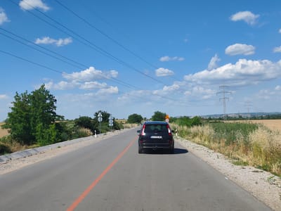 This media file captures a daytime scene on a paved road in Krushovitsa, Bulgaria. The perspective is from a vehicle traveling on the road, looking ahead. The main subject is a black Ford C-Max car, with the license plate "ER 6906 ME," driving in the same direction. The road has a clear orange line painted along its center.

The setting is rural, with dry, tall grasses and some vegetation lining the sides of the road. In the distance, there are fields of what appears to be harvested crops, and further back, industrial structures and power lines are visible. The sky is a bright blue with scattered white cumulus clouds, suggesting fair weather. A yellow triangular road sign with a black border and the number "50" inside is visible on the right side of the road, indicating a speed limit. Several traffic signs and utility poles are also present along the roadside. The overall mood is one of travel through a sunlit, open landscape.