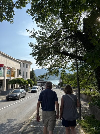 The image shows a view of a street in Bled, Slovenia, taken from the sidewalk. The location is at 46.368 latitude and 14.11 longitude. Two adults, a man and a woman, are walking on the sidewalk in the foreground. The man is wearing a blue polo shirt, khaki shorts, and black sneakers. The woman is wearing a white and black patterned dress with black shorts and white sneakers. They are walking in the same direction, the man slightly ahead of the woman. In the background, there is a busy street with parked cars and people walking and cycling. There is a building on the left side of the image, with a red sign that says "TOTO" in white letters. There is a mountain in the distance, behind the street. The sky is blue with a few white clouds. The trees are in full bloom and the leaves are green. The weather is sunny and pleasant. The image is taken from a low angle, looking up at the street. The image is in focus and the colors are vibrant.
