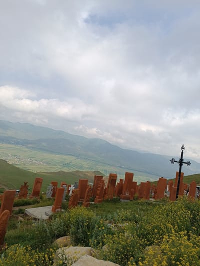 In Hartagyugh, Armenia, this image captures a cemetery on a grassy hillside under a cloudy sky. Rows of upright, reddish-brown gravestones, some shaped like tall rectangular slabs and others with carved details, are scattered across the landscape. Yellow wildflowers bloom in abundance throughout the foreground and between the graves. In the middle ground, a large, black, ornate cross stands prominently to the right. Behind the cemetery, a valley unfolds with a village nestled amongst patchwork fields, and further in the distance, rolling, misty mountains rise up to the horizon. Several figures are visible walking amongst the gravestones in the mid-to-background.