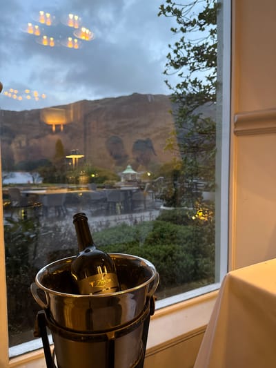 A slightly canted, medium-shot depicts a bottle of red wine chilling in an ice bucket situated on a window sill. The label on the wine bottle is partly obscured but reads "Amor". Through the window, a blurred landscape is visible, including a mountain, greenery, and artificial lights suggestive of a built environment. This is in the city of Glenridding in the United Kingdom. The lighting appears to be subdued, with a focus on the wine bottle and the view outside. A portion of a white tablecloth is visible in the upper right corner.