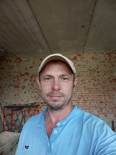 The image is a selfie of a man wearing a light blue collared shirt and a light-colored baseball cap. He has short brown hair, a light beard, and is looking at the camera. He is wearing a black wireless earbud in his right ear. The background consists of a brick wall and a concrete ceiling. In the lower left corner, a wheelbarrow and some red scaffolding with a ladder are partially visible. The scene appears to be an unfinished or under-construction building. The image does not contain any explicit information that can link it to Saratovskaya, Russia.
