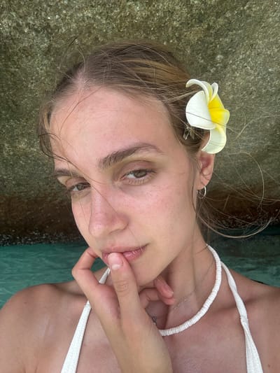 A young woman is pictured in what appears to be a coastal area, possibly in the city of Lam Kaen, Thailand, although no explicit landmarks confirm this. She is wearing a white top and has a white and yellow flower tucked behind her ear. Her hand is near her face, touching her lip. The background includes a rocky surface on one side and greenish water on the other. She is fair-skinned, and appears to be gazing slightly downward. A thin silver necklace is visible.
