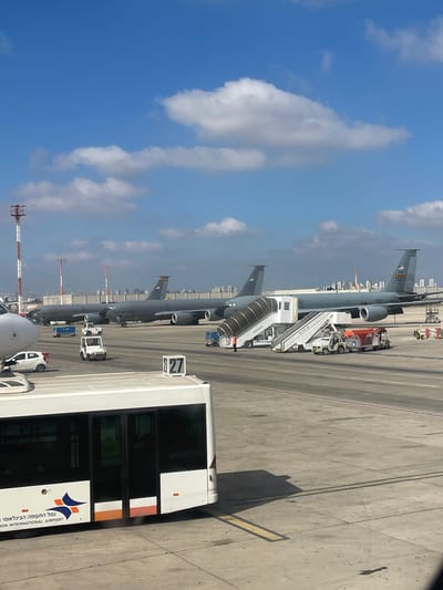 The image depicts an aerial view of Ben Gurion International Airport in Yagel, Israel. The scene is dominated by several large gray U.S. Air Force aircraft parked on the tarmac. In the foreground, a white airport bus is visible, with the text "BEN GURION INTERNATIONAL AIRPORT" and an orange and blue logo printed on its side. Two smaller white utility vehicles are positioned near the aircraft, one of which appears to be a tug. A mobile staircase is attached to one of the aircraft, and a person in an orange vest is standing near it. In the background, a city skyline is visible under a bright blue sky with scattered white clouds. A tall red and white striped tower is also present on the left side of the image. The overall impression is a busy airport environment on a clear day.