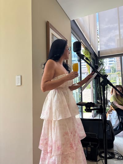 In a well-lit indoor setting, a woman with long dark hair and glasses is dressed in a strapless, floor-length gown adorned with a pink floral pattern. She stands in front of a microphone on a stand, holding a piece of paper in her left hand and a glass of orange liquid in her right. Behind her, a large window offers a view of a green, leafy plant and buildings, suggesting an urban environment. Sunlight streams in, illuminating the scene. Another person, partially visible, is seated behind the woman. Various pieces of audio equipment, including speakers and a case, are positioned nearby. The woman appears focused on the paper she is holding, possibly reading or reciting. The overall atmosphere seems to be that of a performance or a recording session.