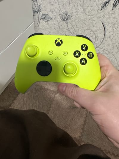 A neon yellow Xbox Series X/S controller is held by a person's right hand in the foreground, with the controller positioned at an angle. The controller is the primary subject, with its buttons and joysticks clearly visible. In the background, there is a grey wall with a floral wallpaper pattern and a white surface, possibly furniture, to the left. The bottom portion of the image shows a furry, brown surface, likely a rug or blanket. The scene appears to be indoors, in a domestic setting. The location is identified as Donskoy, Russia.