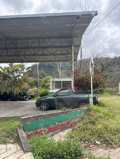 The image shows a partially covered area in Jilotepec, Mexico, potentially a parking or storage space. The structure has a corrugated metal roof supported by a steel frame. Outside the covered area, there's a green pickup truck parked alongside a concrete curb painted with green and red stripes. A pile of tires is stacked near the truck, close to palm trees. The background features lush green foliage, likely part of the mountainous terrain surrounding Jilotepec. The sky is overcast. No people are visible in the frame.