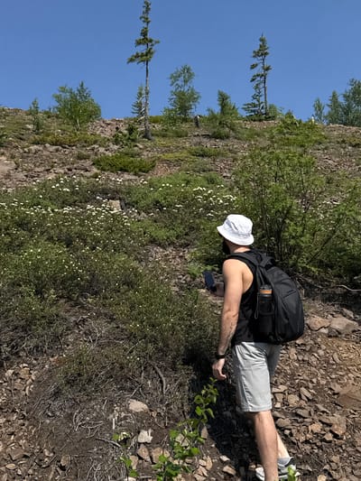 The image shows a person from behind, wearing a white bucket hat, a black t-shirt, light gray shorts, and a black backpack. The person is looking down at a smartphone in their hand. They are on a rocky, uneven terrain with sparse vegetation, including bushes with small white flowers and some trees. The scene appears to be a natural outdoor environment, possibly a hiking trail or a hillside. The lighting suggests it is daytime, with bright sunlight casting shadows. The sky is clear and blue. The overall impression is of a person taking a break during an outdoor activity, perhaps navigating or checking something on their phone. The location is identified as Talnakh, Russia. No visible text is present in the image.