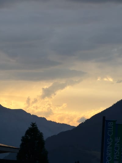 The image shows a sunset over the mountains in Saint-Gervais-les-Bains, France. The sky is filled with dramatic clouds, illuminated by the warm glow of the setting sun. Silhouetted against the vibrant sky are the dark outlines of the mountains and a tall evergreen tree in the foreground. On the right side of the image, two vertical banners are partially visible, displaying text that appears to be in French, with "SPORTING-CLUB" and "PISCINE" being legible. The overall atmosphere is serene and majestic, capturing the natural beauty of the French Alps at dusk.