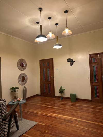 The image shows an interior room with hardwood floors and light yellow walls. A set of three modern pendant lights hangs from the ceiling. Two decorative straw-like mirrors are on the wall along with a vase. A small round table sits near the mirrors with a potted plant. The wall is also decorated with an African-shaped art piece. Two wooden doors with colorful patterns are visible, one near the ceiling and one at floor level. A green trash can and a potted plant sit in the corner of the room.