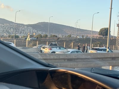A number of vehicles, including a white SUV, are parked on the side of a road. Several people, some appearing to be men, are standing near the vehicles. The scene is set against a backdrop of a city built on a hillside, with mountains in the distance. The sky is clear and a warm light suggests it is either sunrise or sunset. The area appears to be in Turan, Israel, indicated by the landscape and architectural style of the buildings on the hillside. The presence of multiple vehicles and people suggests a possible gathering or event.