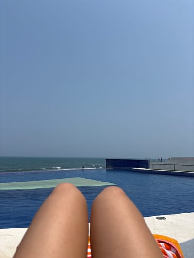 The image, taken in Boca del Río, Mexico, shows a view of an infinity pool merging with the ocean under a clear, light blue sky. In the foreground, a person's legs are visible, with an orange and white striped towel nearby. The pool's edge is tiled in dark blue. The pool appears to blend seamlessly with the sea in the background.