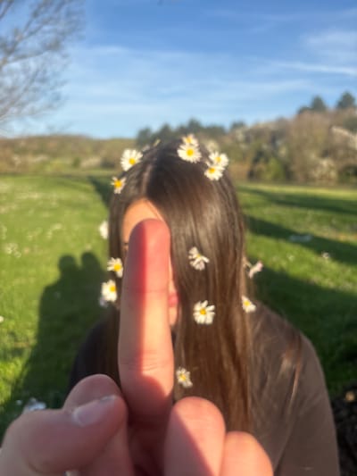 The image is of a person lying in a grassy field, possibly in Beli Brod, Bulgaria, although there are no elements confirming it. The person has brown hair decorated with small white daisies. They are giving the middle finger to the camera. The background includes green grass, trees, and a blue sky. The image is slightly blurry.