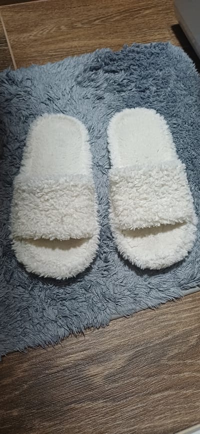 A pair of white, fluffy slippers are displayed on a grey, plush rug, which is situated on a wooden floor. The slippers are oriented side-by-side, with their open fronts facing upwards. The rug exhibits a shaggy texture, and the wooden floor has a visible grain pattern. The image is taken from a top-down perspective, focusing on the slippers and the immediate surrounding rug. There are no people or animals present in the image. The scene appears to be indoors. There is no information available to link this image to the city of Zuevka, Russia, or any specific urban location.