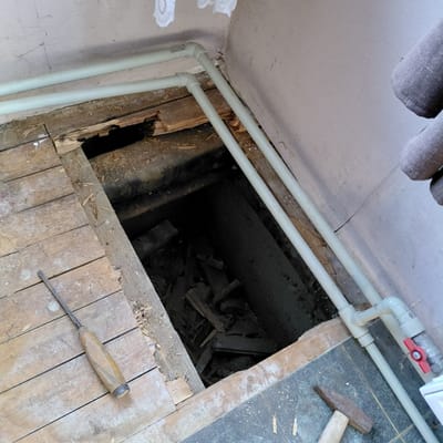 The image depicts a section of damaged wooden floorboards with a dark opening in the center, revealing debris below. Two light-colored pipes run horizontally above the floorboards, with one pipe having a red valve attached to it. A chisel lies on the wooden floorboards to the left of the opening, and a hammer is partially visible in the lower right corner of the image. The surrounding environment appears to be an indoor space with textured walls and some white material hanging from the ceiling. The scene suggests ongoing repair or demolition work within a residential or utility area. Based on the provided context, this scene is located in Skuķīši, Latvia.