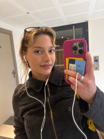 A young female, identified as the subject, is centered in a close-up, reflected image, likely a mirror selfie, within an indoor environment in Nogent-sur-Marne, France. She possesses fair skin, light brown wavy hair styled with bangs, and visible freckles on her nose and cheeks. Black-framed glasses are pushed onto the top of her head. She wears white wired earbuds in both ears, with the cord extending down the front of her torso. Her attire includes a dark, possibly black or charcoal, denim jacket with a brass-toned zipper fastened to the neck.
Her left hand, positioned towards the viewer, holds a bright pink smartphone, which features a triple-lens camera array consistent with an iPhone model. Tucked into the back of the phone's case are two cards: a yellow card partially visible behind a blue and white French identification card. The visible French ID card displays a photographic portrait of a female, inverted text including "MINISTÈRE DE L'INTÉRIEUR" and "TITRE DE SÉJOUR," indicating a French residence permit. The subject's left hand bears two rings: a wide, patterned band on her ring finger and a thinner band on her pinky finger.
The reflected background elements include a white acoustic drop ceiling with square perforated panels. To the left of the subject, a light-colored door with a dark handle is visible. To the right, behind the phone, an intercom panel with buttons and a numerical keypad is reflected on a wall, adjacent to a dark doorframe. A checkered floor pattern is subtly reflected in the lower left quadrant of the image.