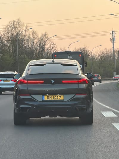 The image depicts a dark grey BMW X6 (Luxembourg registration L DN 1417) from a rear perspective, positioned in traffic on a multi-lane road in Foetz, Luxembourg. Its taillights are illuminated. Immediately ahead is a dark grey bus displaying "508" on its rear. To the left of the X6, a silver BMW SUV (Luxembourg registration L BM 2210) is also visible. Several other vehicles are further down the road. The road surface is asphalt with white lane markings, including broken lines and rectangular patterns on the right shoulder. The background features bare deciduous trees, multiple streetlights, and a cellular communication tower. The sky exhibits an orange and pink hue, indicative of sunrise or sunset conditions. All vehicles appear to be stationary or moving slowly in a traffic queue.