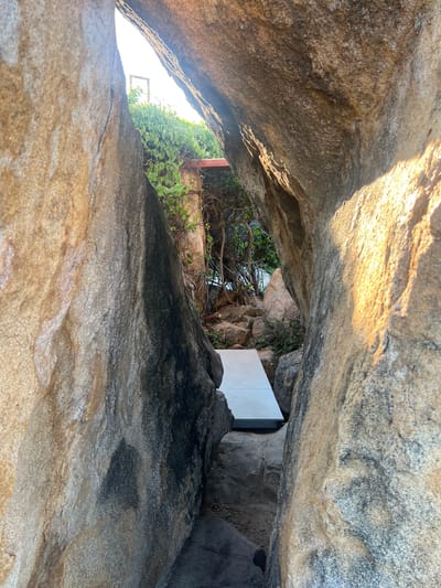 A pathway of light-colored concrete slabs leads through a narrow passage between large, weathered boulders. The rocks display a rough, textured surface with varying shades of brown, gray, and black. Lush green foliage, with vines clinging to branches, grows in the background, partially obscuring a distant structure with a roof. The scene is illuminated by natural light, casting shadows and highlighting the contours of the rocks. The location appears to be an outdoor environment, possibly a garden or a natural landscape feature within a developed area, in Vĩnh Hải, Vietnam.