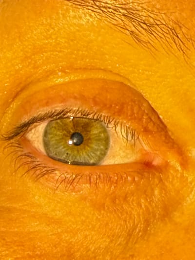 An extreme close-up image captures a human eye in a rotated, vertical orientation. The iris displays a striking hazel-green color, interspersed with warmer brown tones and intricate radial patterns. The central pupil is black and prominently reflects two distinct points of bright light, suggesting specific artificial light sources. Dark, fine eyelashes are visible along both the upper and lower eyelids.
A segment of a dark eyebrow is partially visible along the left side of the frame. The surrounding skin, including the eyelids and adjacent facial areas, exhibits a warm, yellowish-orange hue. The close proximity of the shot highlights the skin's detailed texture, including fine lines, wrinkles, and pores across the entire visible surface. The lighting appears direct and strong, creating subtle sheen on the skin and the clear reflections within the pupil. There is no discernible activity taking place, and no text is visible within the image. The image itself does not provide specific visual cues to its location in Leicester, United Kingdom.
