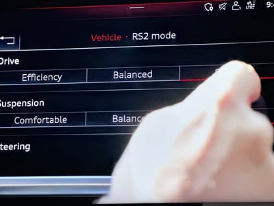 The image shows a car's infotainment screen displaying various driving modes. The "Vehicle - RS2 mode" is prominently displayed in red text. Below this, the screen shows categories such as "Drive," "Suspension," and "Steering." Under "Drive," the options "Efficiency" and "Balanced" are visible. Under "Suspension," "Comfortable" and "Balance" are visible. A blurred hand is interacting with the screen, touching the "Balanced" option for the suspension. The background is dark, consistent with a car's interior at night or in a dim environment. There is no visible indication of the city of Bezden, Bulgaria.