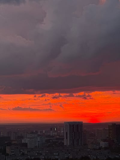 The image depicts a dramatic sunset over the city of Savenki, Russia. The sky is ablaze with vibrant hues of orange and red, transitioning to a deep, ominous purple and gray in the upper portion, indicating a significant cloud cover. The setting sun is visible as a bright, concentrated orb just above the horizon line, casting a warm glow. Below the sky, a dense urban landscape fills the frame, characterized by a variety of buildings. Prominent among these are several multi-story residential blocks and a taller, modern skyscraper with a distinctive facade. The architecture suggests a mix of older and newer construction. The city is viewed from an elevated perspective, allowing for a panoramic sweep of the urban sprawl. The overall impression is one of a powerful natural phenomenon contrasting with the static, man-made environment of the city.