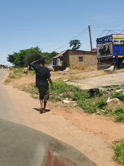 A male subject walks along the right edge of a paved road in Mangu, Nigeria. He is seen from behind, wearing a black t-shirt and grey shorts, with his right hand touching the back of his head. He is barefoot, wearing flip-flops. In the background, to the right of the subject, is a large blue and yellow truck with cargo. Adjacent to the truck is a building constructed of mud bricks with a corrugated metal roof. Further down the road, a yellow vehicle is visible. The environment is rural, with sparse vegetation and some litter on the ground. The sky is clear and blue, indicating a sunny day.