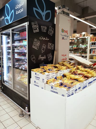 The image shows a retail environment, likely a supermarket, with a focus on a display of cookies and a refrigerated display case. In the foreground, stacked boxes of "J'aime Cookies" are prominently displayed. These boxes are predominantly yellow and blue, with Arabic text visible, indicating "Cookies" and "20 Pieces." The cookies themselves are packaged in yellow wrappers with blue accents.
To the left of the cookie display is a large, black, refrigerated display case with glass doors. The interior of the case is filled with various frozen food items, including what appear to be bags of frozen vegetables or pastries. The exterior of the refrigerator features Arabic text, including "منتوجات مجمدة" (Frozen Products) and "Nuggets," along with stylized graphics of frozen foods like ice cream and burgers.
In the background, shelves stocked with various packaged goods, possibly spices or dry goods, are visible. Above the refrigerated case, a blue sign with stylized plant-like graphics displays more Arabic text. To the right of the refrigerated case, a tall, white vertical display unit has a logo with Arabic text that appears to say "mg" and "yufalik kul yom akmar min 140" (likely a promotional slogan). Several cans are stacked on top of this unit.
The scene is brightly lit, suggesting it's daytime. The floor is tiled with light-colored squares. There are no people visible in the image. The overall impression is that of a well-stocked grocery store aisle, with a focus on promotional displays.