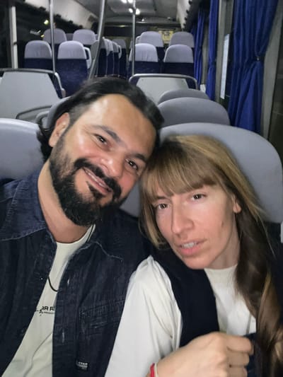 A couple is taking a selfie on a bus. The man on the left has dark hair and a beard and is wearing a denim jacket over a white t-shirt. The woman on the right has long brown hair and bangs and is wearing a black vest over a white t-shirt. The background shows rows of empty seats on the bus, with blue curtains on the right. The bus appears to be in a stationary position, possibly at a bus stop or terminal in Santa Perpètua de Mogoda, Spain, as indicated by the context. The lighting is dim, suggesting it might be evening or nighttime.