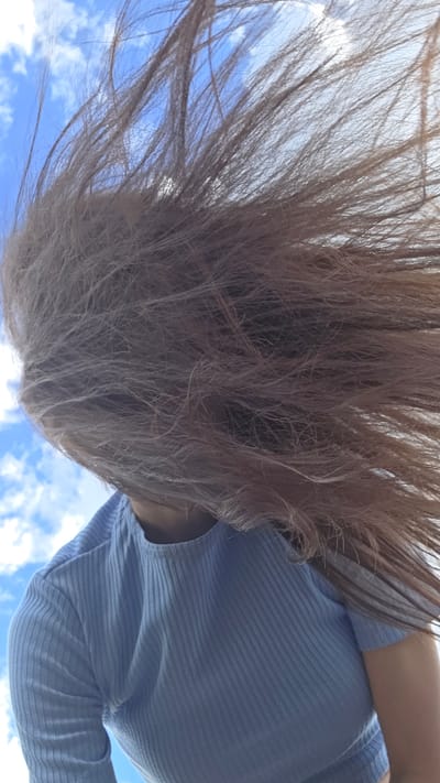 A person's head and shoulders are visible from a low-angle perspective, with their long, light brown hair blurred and blowing in the wind across the frame. The person is wearing a light blue, ribbed, long-sleeved shirt. The background consists of a bright blue sky with scattered white clouds. There is no discernible city or specific location information visible in the image.