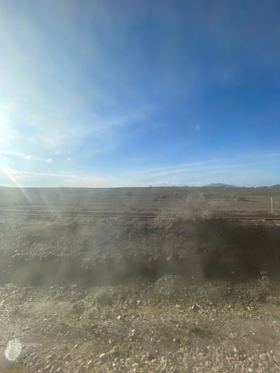 The image presents a wide, arid landscape under a bright, clear blue sky, likely captured from a moving perspective, such as a train or car. The top half of the frame shows a vast expanse of blue sky with scattered, delicate white clouds. A prominent sun glare or lens flare emanates from the lower right portion of the sky, indicating a bright sun and possibly a window separating the camera from the scene.
The lower half of the image features a dry, rocky, and sparsely vegetated terrain. The ground is covered with what appears to be gravel, dust, and low-growing, scrub-like bushes and plants, typical of a semi-desert environment. Running horizontally across the middle ground are two distinct parallel lines, strongly suggesting a railway track traversing the landscape. In the distant background, along the horizon, faint, hazy outlines of mountains or hills are visible. There are no people or discernible man-made structures other than the potential railway line. The scene depicts a sunny day with no visible adverse weather conditions. The overall impression is one of vast, open wilderness, consistent with the location context of Jbilate, Morocco. No visible text is present.