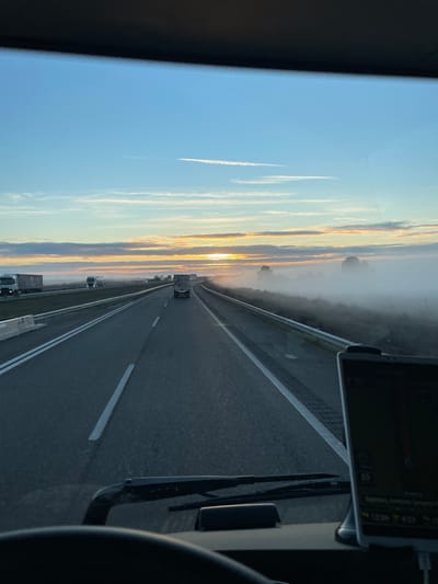 A highway stretches ahead under a sky painted with sunrise colors. The road, lined with white lane markers and a roadside barrier, is traveled by a few trucks. Wisps of clouds hang low, blending with a distant hazy horizon. A phone displaying navigation details is mounted on the dashboard, along with a windshield wiper. The view is from inside a vehicle, capturing a roadside scene at dawn or dusk.