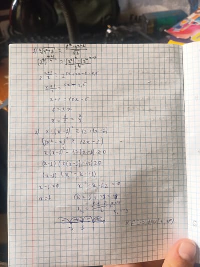 The image shows a page from a squared notebook, likely a math notebook, filled with handwritten mathematical equations and inequalities. The handwriting is in dark ink, possibly a pen. The top left corner of the page has a faint handwritten number "1)" followed by a mathematical expression. Below this, there are several lines of mathematical calculations and transformations. Further down, a "2)" marks the beginning of another problem, which appears to be an inequality. This second problem involves algebraic manipulation and leads to a quadratic equation. The calculations include finding roots of the quadratic equation and analyzing intervals on a number line. The bottom right corner of the page has a handwritten note or symbol. The notebook page itself is lightly gridded with blue lines. The background appears to be a surface with some texture, possibly a desk or table, with a blurry, out-of-focus object to the left that could be a lamp or electronic device. The lighting in the scene is somewhat dim, casting shadows. The overall context suggests someone is working on math problems. There is no information within the image that relates to the city of Kyzyltog (Krasnogorsk), Uzbekistan.
