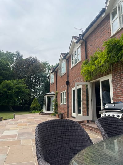 A two-story red brick house is captured from an outdoor patio perspective. The house features multiple white-framed windows and white double doors. A green bamboo plant is near the roof, and climbing vines adorn a bay window on the ground floor. A patio set with woven chairs and a glass-topped table is in the foreground. A grill is visible near the doors. Green trees and lawn complete the scene.