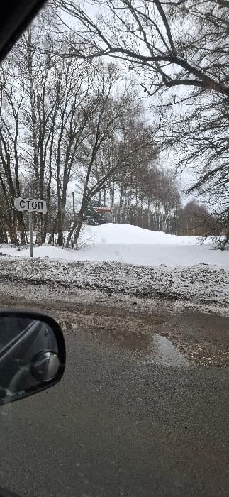The image depicts an outdoor winter scene in Shornovo, Russia, viewed from a vehicle. The immediate foreground shows a dark asphalt road surface with puddles and scattered gravel. To the lower left, a portion of a black vehicle side mirror is visible, incorporating a small convex spot mirror. A dirty snowbank lines the edge of the road, behind which a white rectangular sign with the Cyrillic word "СТОП" (STOP) in black lettering is mounted on a pole. Further back, a significant snow-covered mound occupies the midground. Behind this mound, a structure with a reddish-orange roof is partially visible. The background consists of numerous bare, deciduous trees under an overcast sky, suggesting late autumn or winter conditions. No individuals are present in the frame.