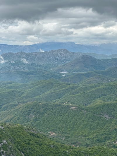 The image depicts a mountainous landscape in Brskut, Montenegro. The scene is dominated by a series of densely forested mountain ridges, with varying shades of green indicating different vegetation types. The ridges are layered and extend into the distance. Some small buildings, likely residential, are sparsely distributed on the slopes. A river cuts through one of the valleys. The sky is filled with heavy, gray clouds, casting a diffused light over the scene. The image is taken from an elevated position, providing a panoramic view of the mountainous terrain.