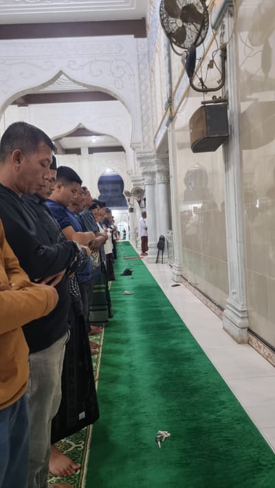 A group of Muslim men are standing in prayer on a green carpet inside a mosque. They are aligned in rows, facing the same direction. The mosque appears to be in the city of Kambam, Indonesia, as indicated by the presence of Islamic architectural elements and the cultural context. The men are dressed in traditional Islamic attire and casual wear. The environment is well-lit, with white walls and decorative arches. A ceiling fan is visible on the wall, along with some electrical wiring. Some of the men are wearing sarongs, a common garment in Indonesia. The men are engaged in prayer, with their hands folded. A set of keys is visible on the carpet in the foreground. The overall scene depicts a religious gathering in a mosque.