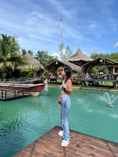 A young woman stands on a wooden pier next to a body of turquoise water. She is wearing ripped blue jeans and a gray tank top, with a small orange purse slung over her shoulder. Her hair is dark and falls to her shoulders, and she appears to be shielding her eyes from the sun, suggesting a bright, sunny day. The water features a fountain in the distance.
Behind the woman are several structures with thatched roofs, reminiscent of traditional Southeast Asian architecture. Lush green palm trees and other tropical foliage surround the buildings. A long, wooden boat with colorful trim is docked nearby, tied to the pier. Further in the background, a tall, red and white communication tower rises against a bright blue sky with scattered white clouds.
The scene appears to be a tourist destination, possibly a resort or a recreational area, given the water features, boat, and unique architecture. The time of day is clearly daytime, indicated by the bright sunlight and clear sky. The overall atmosphere seems relaxed and picturesque, capturing a moment of leisure in a tropical setting. The location cues point to Tambon Pa Kae Bo Hin, Thailand.