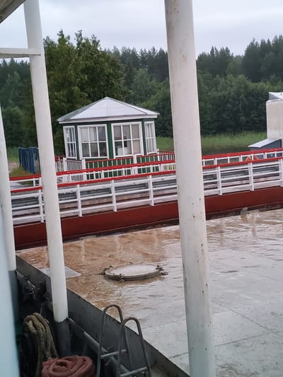 The image shows a ferryboat docked on a body of water. The ferryboat has a red hull and a white railing with red accents. On the deck, there is a small white building with green trim and multiple windows, possibly a control booth. A round metal hatch is visible on the wet deck. In the background, a dense green forest lines the shore. The sky is overcast. The scene is consistent with the environment in Belousovo, Russia, a town located near rivers and forests.