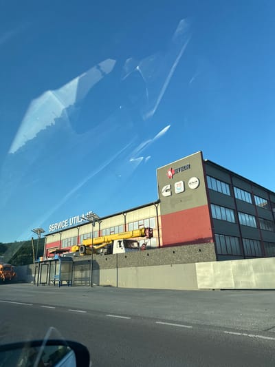 The image depicts a commercial building with the prominent signage "SERVICE UTILAJ" in white lettering against a blue sky. To the right, another section of the building displays logos, including "METEOR", "Cummins", and circular emblems. A yellow crane is positioned in front of the building. In the foreground, a bus stop with a blue bench and glass enclosures is visible. An orange industrial vehicle is parked to the left of the bus stop. The scene appears to be located along a road in or near Trușeni, Moldova, given the presence of the bus stop and commercial structure. The sky is clear blue with some reflections or streaks, possibly from a window or camera lens.