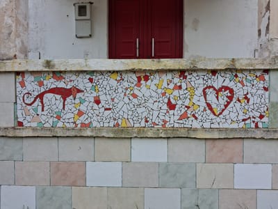 The image shows a portion of a building facade. The upper part of the frame contains a white wall with peeling paint. At the top center of the image, there is a pair of closed, dark red doors with metal handles. Beneath this, a decorative tiled panel features a mosaic design. The mosaic depicts a red stylized dog or fox-like creature on the left, a scattering of red, yellow, and other colored tiles in the center, and a red heart shape on the right. The tiles appear to be broken and arranged in a pattern. Below the mosaic, a wall is covered in rectangular tiles of varying pastel colors. An electrical box is mounted on the wall above the mosaic and to the left of the doors. This image was taken in Soure, Portugal.