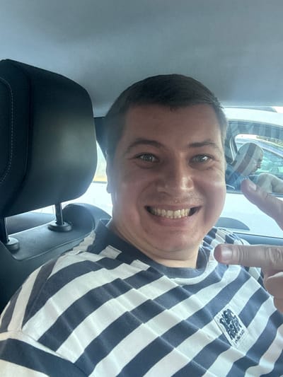 The image shows a selfie of a man in a car, likely in Narbonne, France. He is wearing a striped shirt and smiling. His right index finger is pointing towards the camera. In the background, another person is visible in a car driving alongside. The setting appears to be a sunny day.