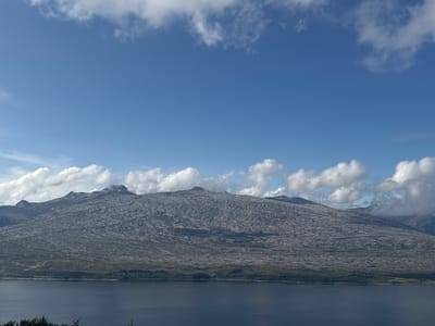 An expansive, clear blue sky dominates the upper half of the image, with scattered white, fluffy clouds drifting across. Below the sky, a vast, rugged mountain range stretches horizontally across the mid-ground. The mountains are covered in a coarse, greyish-brown texture, suggesting rocky terrain with sparse vegetation. The slopes are uneven and weathered, with distinct shadows cast by the sunlight, highlighting the contours of the landscape.

A narrow band of dark green trees and shrubbery forms a border along the base of the mountains. Below this, a large body of dark blue water, likely a fjord or lake, occupies the lower portion of the frame. The water is calm, with only a slight ripple effect visible on the surface. In the far distance, the mountain peaks fade into a hazy, lighter blue, suggesting a considerable distance. The overall scene depicts a natural, mountainous landscape with a body of water under a partly cloudy sky. This scene is consistent with the geography of Flostrand, Norway.