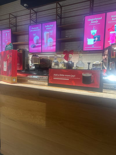 The image shows the interior service counter of a Starbucks coffee shop in Saint-Germain-en-Laye, France, during the winter holiday season. The lower half of the frame is dominated by a light-colored wooden customer counter. Above this, a darker service counter features various items. On the far left, a tall, red Starbucks branded box with gold accents and "Invite the Joy" text is visible. Adjacent to it are components of a commercial coffee machine, including a clear container holding dark roasted coffee beans. In the center, four clear glass bottles with pour spouts, likely containing syrups, are arranged; two hold clear liquid, one dark liquid, and one lighter liquid, with a red bow tied to the second bottle from the left. To the right of the bottles, a prominent red and black framed display sign advertises "Add a little more joy!" and "l'Espresso Christmas Blend," featuring an image of a dark beverage in a clear mug, with accompanying French text. A small white box with blue text is partially visible behind the sign. On the far right, a female employee with dark hair is partially visible behind the counter, looking towards the right out of frame. The background is composed of multiple brightly lit digital menu boards displaying Starbucks seasonal promotions in French, including "The iconic chocolate mix," "Toffee Nut season," and "Gingerbread family." Empty wooden shelves supported by dark metal frames are positioned between and above the menu boards against a white tiled wall. A red bow is also attached to the metal shelving system. A small black security camera is mounted on the ceiling at the top-center. The scene is illuminated by artificial light.