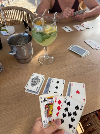 A high-angle, medium shot captures a card game in progress on a wooden table at an outdoor cafe in Port de Pollença, Spain. The scene is dominated by the table, with a partially visible person in the upper background holding a hand of cards. To the left, an ice bucket with tongs and ice sits next to a tall glass filled with a yellow beverage, ice, mint, and lemon slices. In the foreground, several playing cards are spread across the table. These include three Aces of Spades, two Aces of Hearts, a 3 of Spades, a 7 of Hearts, and a 9 of Spades. A hand in the lower right corner is holding a King of Clubs, a 10 of Diamonds, and a 9 of Spades. In the background, a woven chair is visible. The lighting suggests a sunny day.